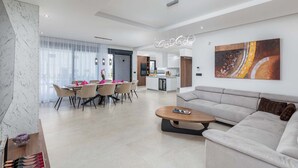 A modern living room with a sofa, a dining area with chairs, a kitchen in the background, and a large painting on the wall. - Villa Ana Skicini in Jursici (Jursici)