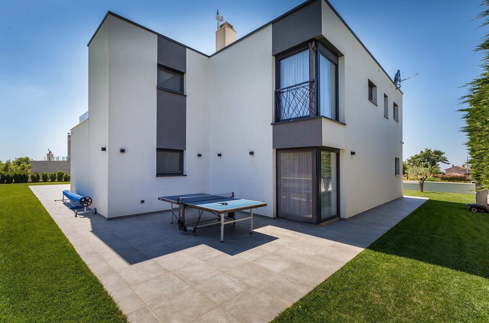A modern two-story house with a flat roof, a tennis court, and a large sliding glass door.