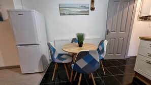 Une cuisine compacte avec un réfrigérateur blanc, une table ronde en bois agrémentée d’une plante en pot, et des chaises à carreaux bleus et blancs.