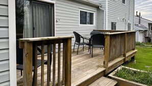 Eine Holzdar Terrasse mit Sitzgelegenheiten, eine Schiebetür und ein weißes Haus mit Giebeldach.