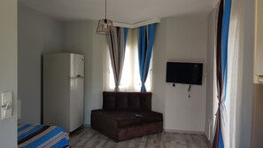 A small, sparsely furnished room with a sofa, a flat-screen TV mounted on the wall, a refrigerator, and a window with curtains. - Taşev Suites (Bodrum)