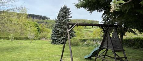 Un conjunto de columpios de madera con un tobogán en un área cubierta de césped, con árboles y una colina al fondo.