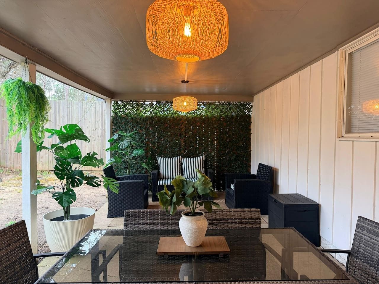 A covered patio area with a glass table, wicker chairs, a hanging light fixture, and a backdrop of greenery.