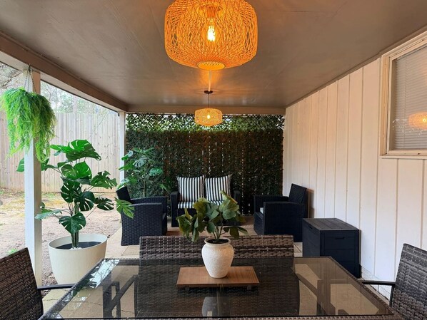 Ein überdachter Patio-Bereich mit Glastisch, Korbstühlen, einer hängenden Leuchte und einem Grünpflanzen-Hintergrund.