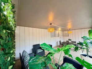 Een kamer met een hoge plafond, witte muren en een plafondventilator. Er staan twee zwarte rieten banken met gestreepte kussens, een eettafel met stoelen en een grote groene plant in het midden.