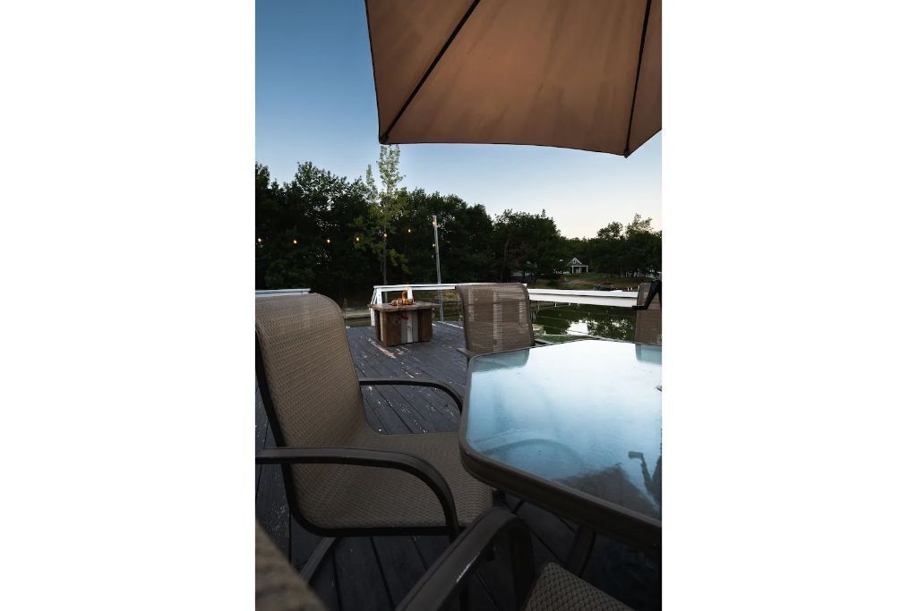 Eine Terrasse im Freien mit Glastisch und -stühlen, ein Holzdeck und Blick auf ein Gewässer und Bäume.