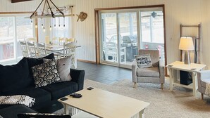 Un vaste salon doté d’un haut plafond, de poutres apparentes, d’une cheminée, d’un coin repas et offrant une vue dégagée grâce à de grandes portes-fenêtres coulissantes.