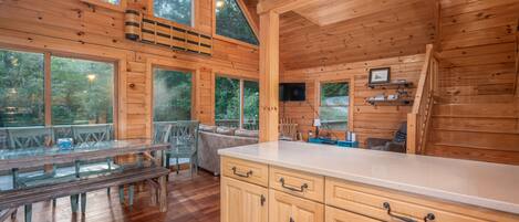 Una cabaña de madera con comedor, cocina y ventanas amplias que ofrecen vistas del bosque circundante.
