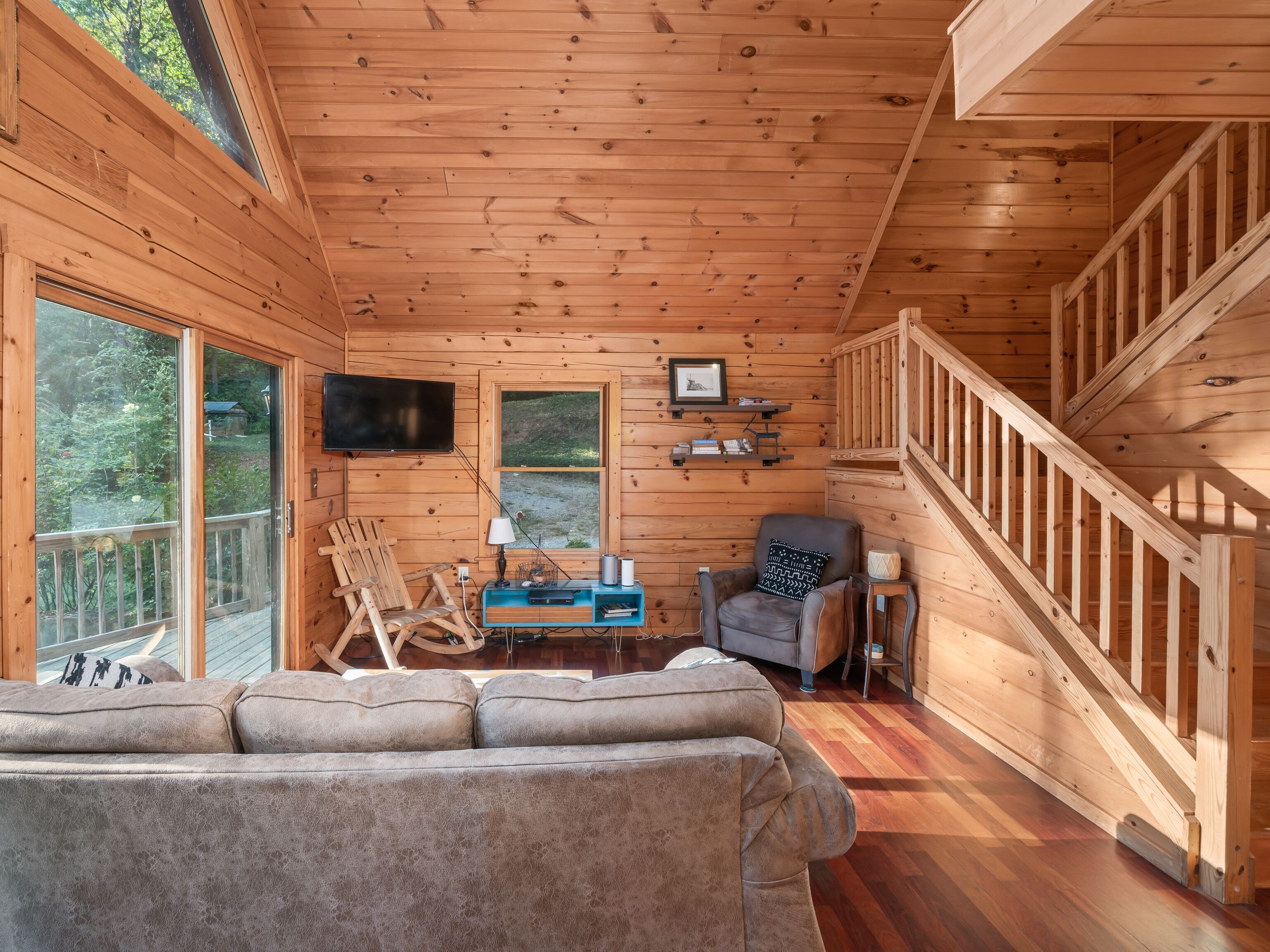 A wooden cabin interior with a sofa, chairs, a TV, and a staircase.
