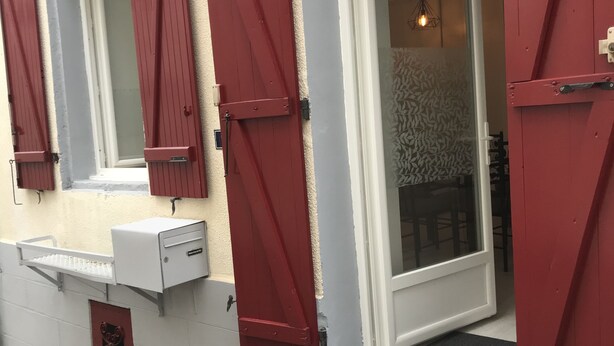 Een gebouw met rode luiken, een witte deur en een klein buiten terras met een bankje.