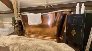 Una vasca da bagno in rame in una stanza con pavimento in legno, un tappeto in pelliccia, una piccola lanterna e un mobiletto in legno scuro con una serratura dorata.
