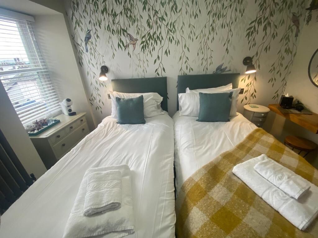 A double room with two beds, each with a pillow and a folded towel. There is a window with blinds, a bedside table with a lamp, and a wall with a leaf pattern.