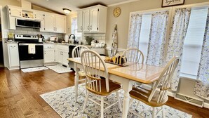 Une cuisine avec des meubles blancs, une table à manger en bois et des chaises. Il y a une horloge au mur et une fenêtre avec des rideaux.