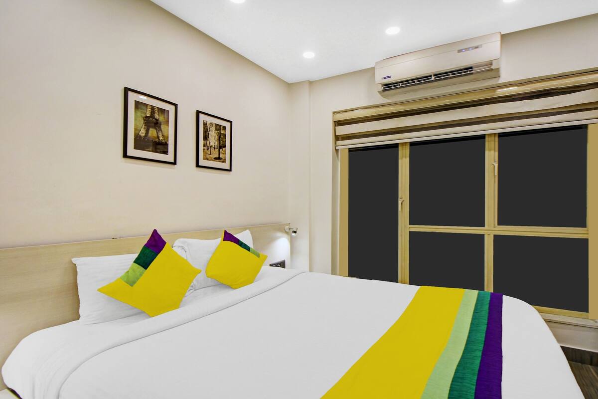 A hotel room with a bed, two framed pictures on the wall, an air conditioning unit, and a window with blinds.