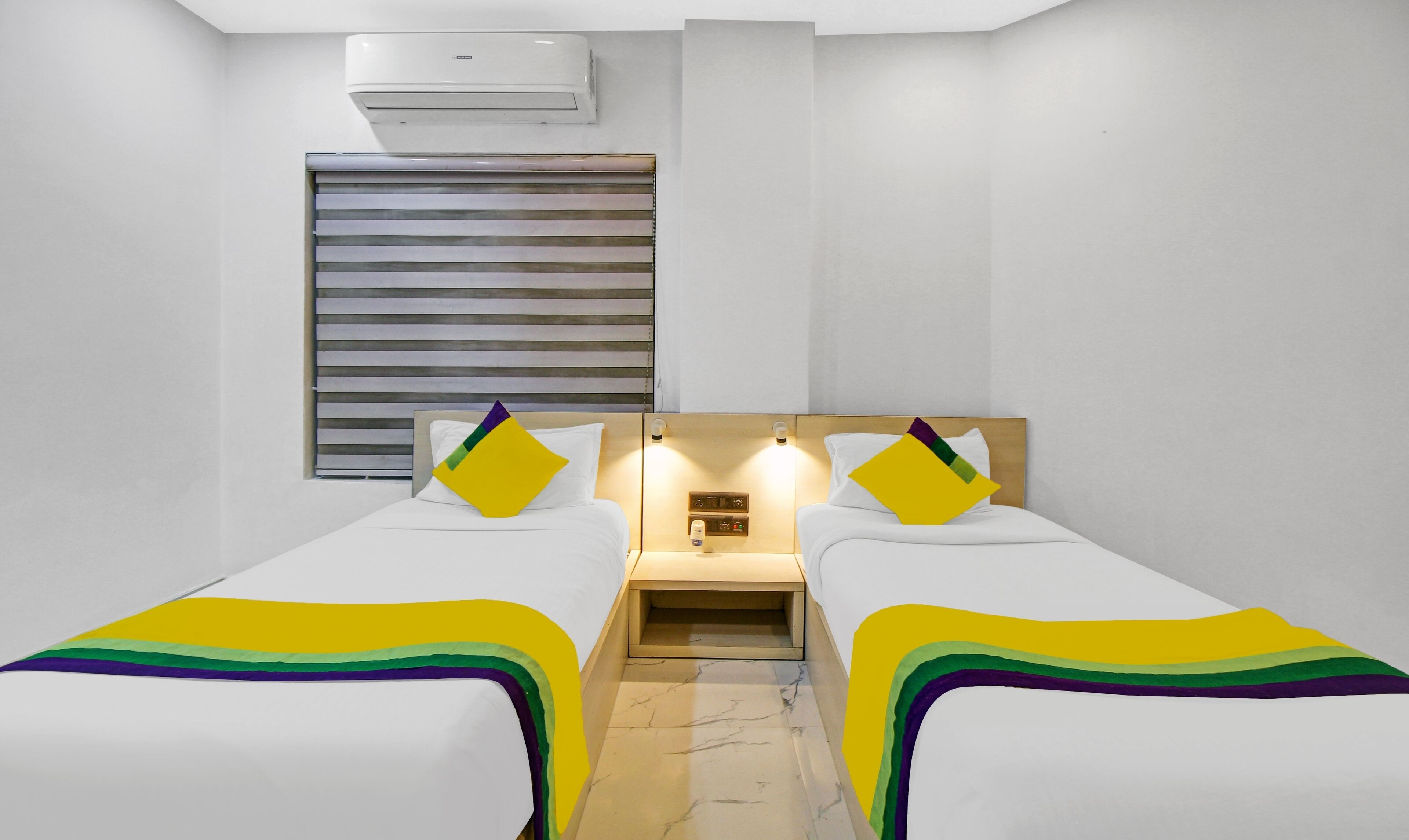 Ein Hotelzimmer mit zwei Einzelbetten, einem Nachttisch mit Lampe, einem Fenster mit Jalousien und einer Klimaanlage an der Wand.