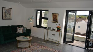 A living room with a green sofa, a round coffee table, a patterned rug, a small side table with a lamp, and a sliding glass door leading to an outdoor balcony.