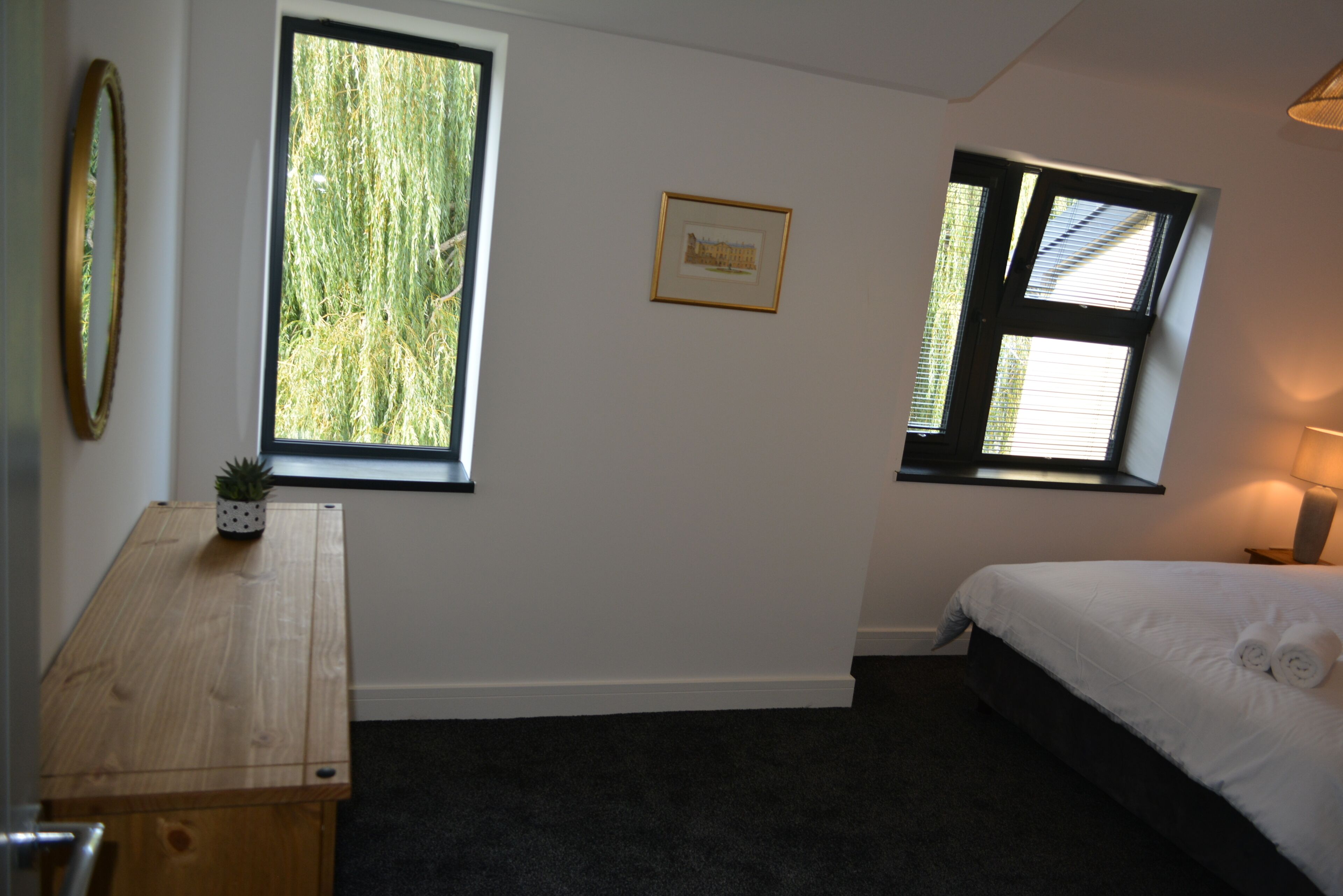 A bedroom with a bed, two windows, a desk with a plant, and a framed picture on the wall.