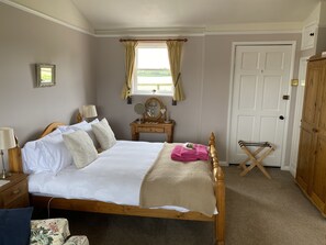 Ein ordentlich bezogenes Bett mit weißen Leinen, ein hölzerner Kopfteil und eine beige Tagesdecke. Ein Nachttisch mit Lampe und ein kleiner Tisch stehen neben dem Bett. Im Eck befindet sich ein Holzschrank, und an der Wand ist ein Fenster mit Vorhängen zu sehen.
