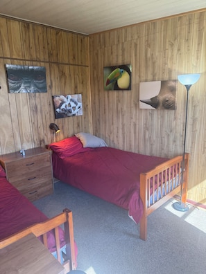 Une pièce aux murs recouverts de lambris en bois, un lit avec une couvre-lit rouge, une table de chevet avec des tiroirs, une lampe et des œuvres d’art accrochées au mur.