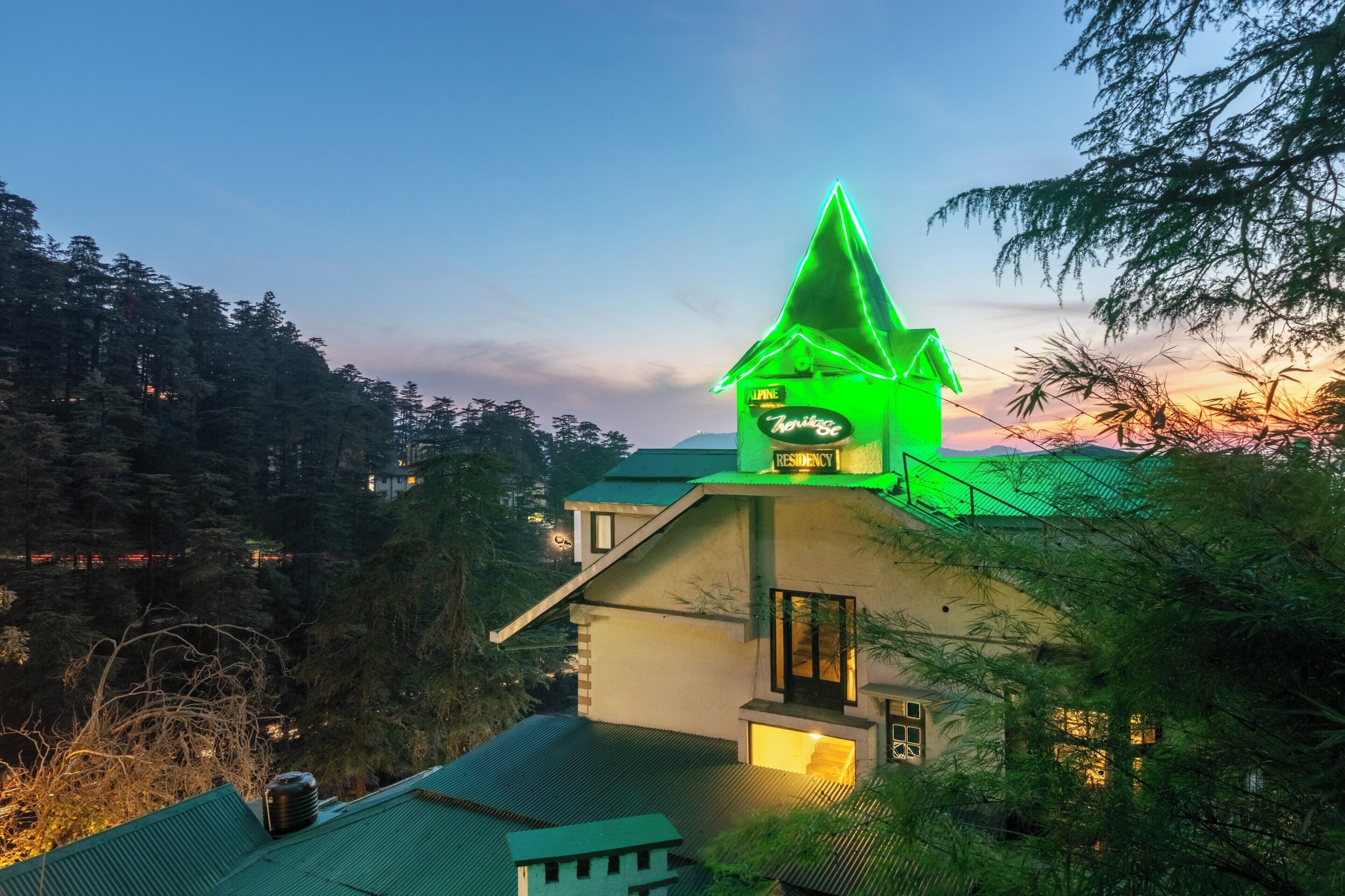Un edificio con una torre iluminada en verde, rodeado de árboles y un atardecer al fondo.