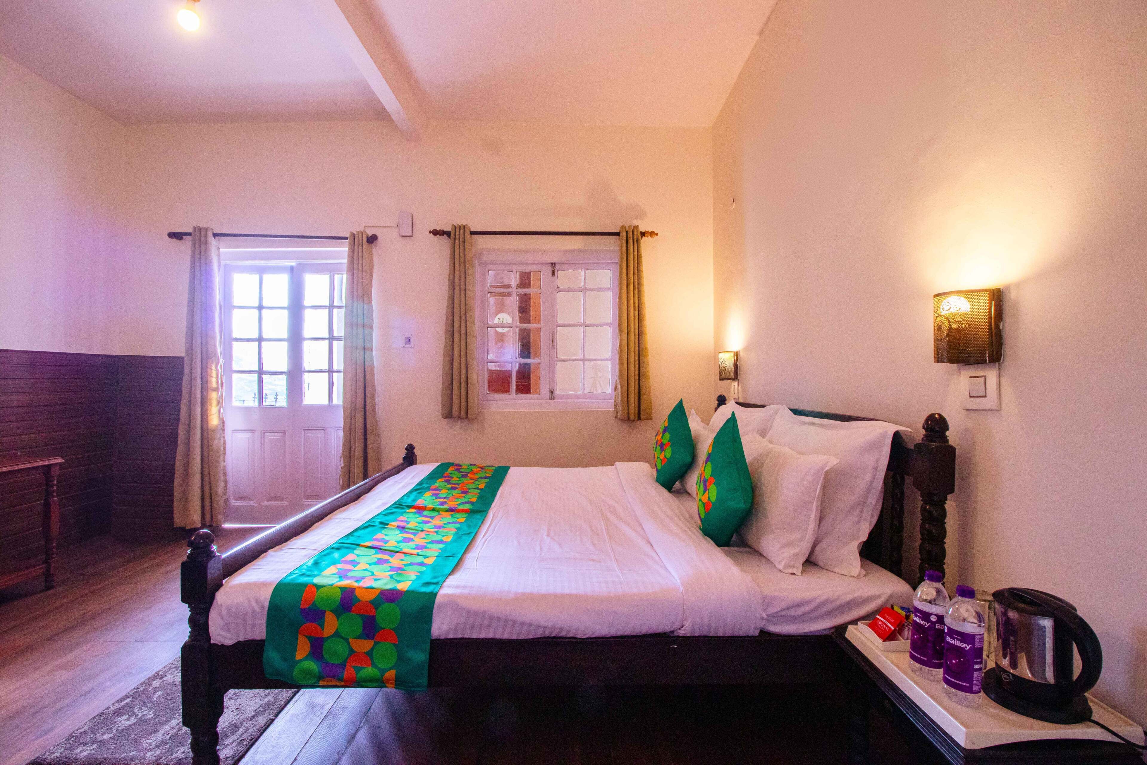Hotelkamer met een bed, gordijnen, een raam, een wandlamp en een nachtkastje met een waterkoker en flessen.