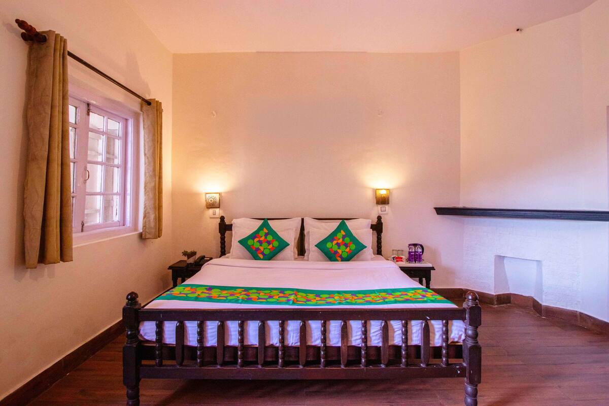 A bed with a patterned bedspread, two wall-mounted lamps, a window with curtains, and a nightstand with a lamp.