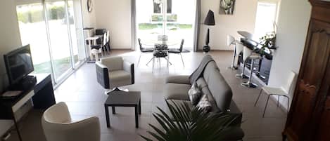 Ein modernes Wohnzimmer mit einer Couch, einem Couchtisch und Sesseln. Es gibt einen Essbereich mit Tisch und Stühlen. Ein Fernseher steht auf einem Schrank, und eine Lampe steht auf einem Tisch.