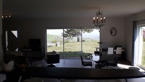En moderne stue med et stort skydedørsvindue, der fører ud til en balkon. Der er et spisebord med stole, et fjernsyn og en lysekrone.