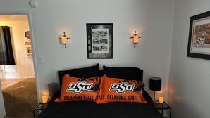 A hotel room with a bed featuring orange and black bedding, two wall-mounted lamps, a framed picture on the wall, and a framed sign on the wall.