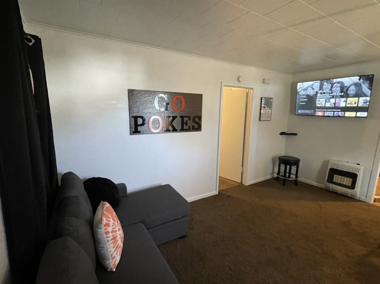 A living room with a grey sofa, a wall-mounted TV, and a sign that reads 'GO POKES'.