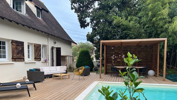 A backyard with a wooden deck, a swimming pool, a covered seating area, and a white house with shutters.