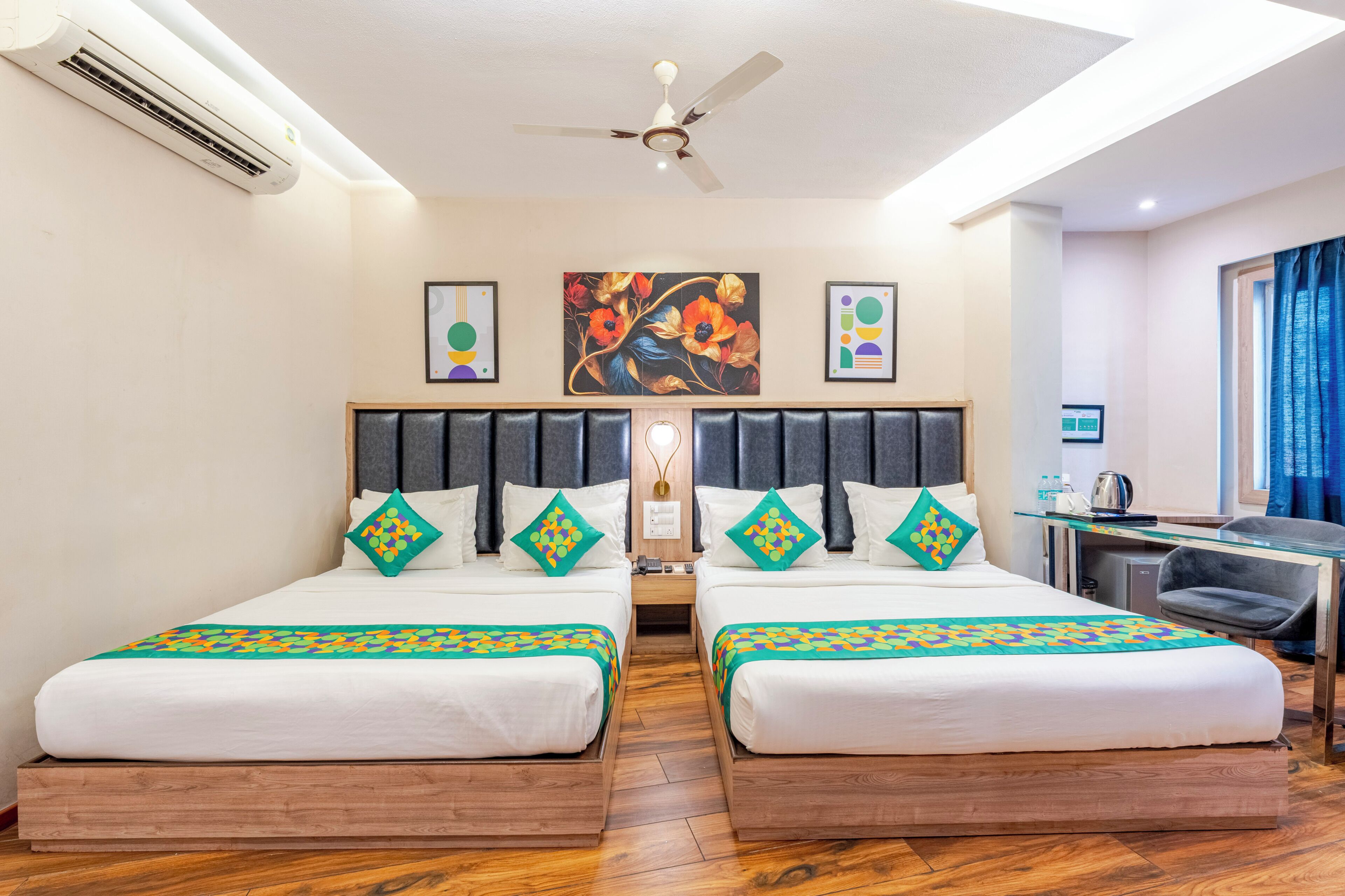 A hotel room with two beds, a desk, a chair, an air conditioner, a ceiling fan, and artwork on the wall.
