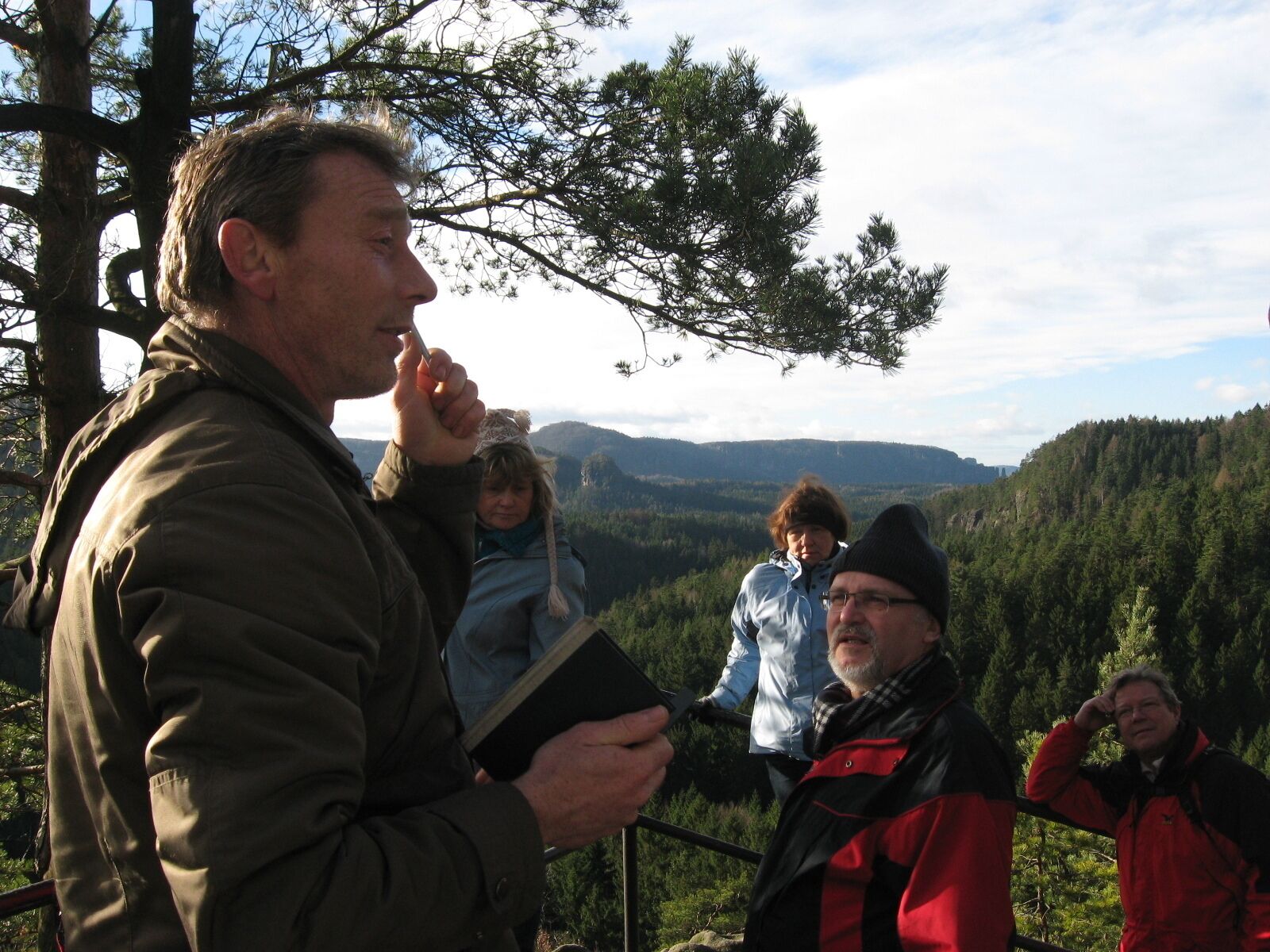 Eine Gruppe von Personen steht draußen, möglicherweise an einem Aussichtspunkt, mit einer bewaldeten Landschaft im Hintergrund.