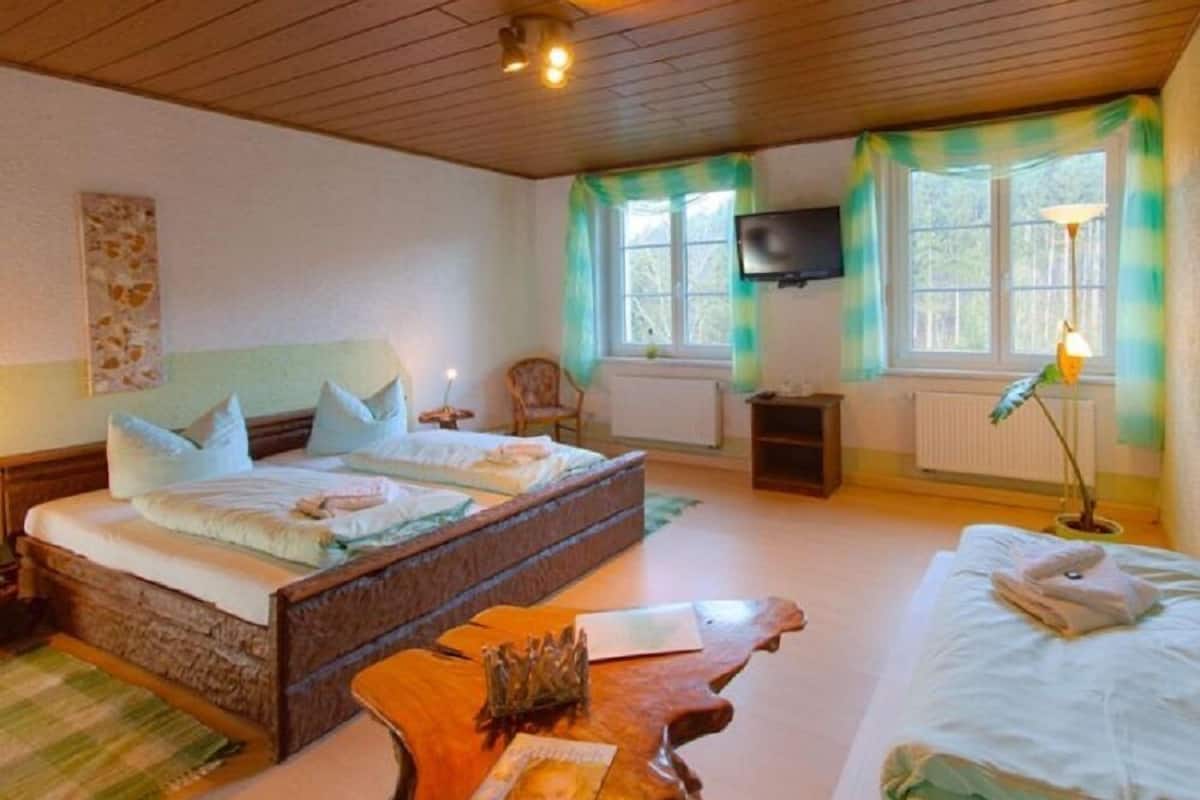 Un dormitorio con una cama grande, cabecera de madera, mesita de noche con una revista, una silla, televisor montado en la pared y ventana con cortinas.