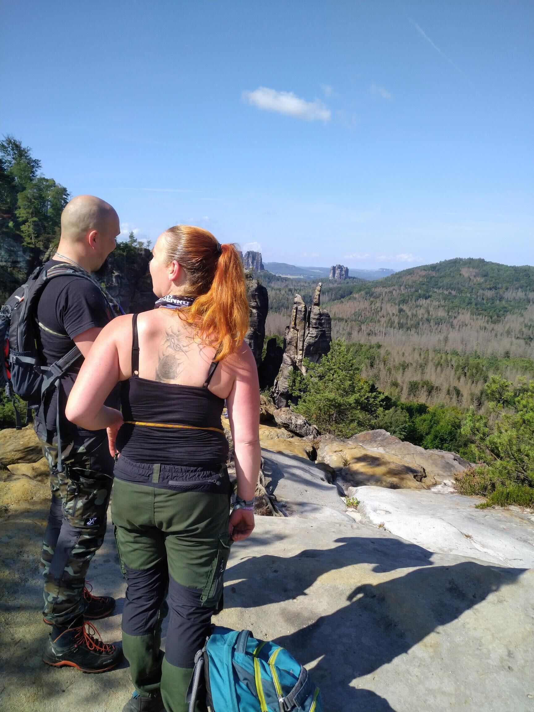 Two people standing on a rocky ledge overlooking a forested valley with a clear sky.