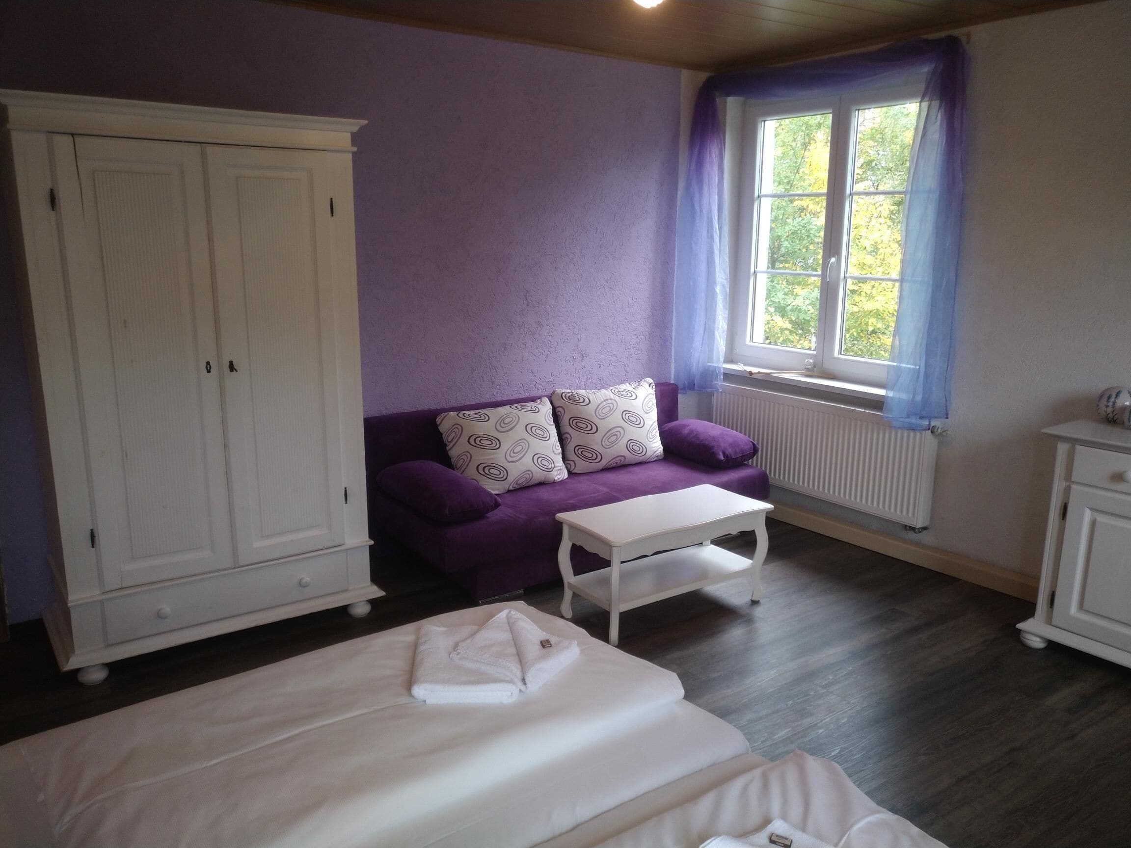 Un dormitorio con una cama, un sofá púrpura, una mesa blanca y un armario.