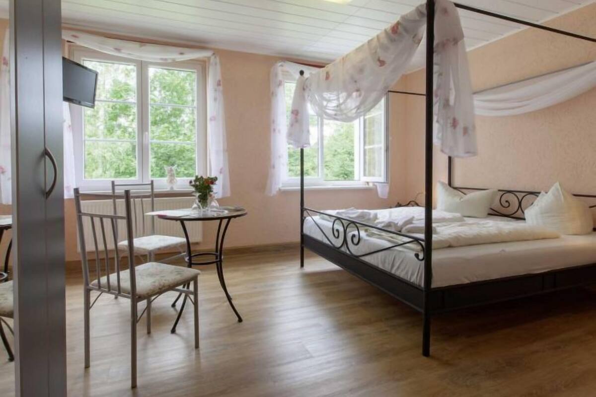 Una cama con dosel, una mesita redonda con un florero, y una silla en una habitación con suelo de madera y ventanas amplias.