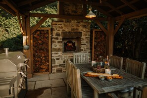 An outdoor patio with a stone fireplace, wooden table, chairs, and a grill. - Rural Holiday Cottage With Log Burner (Buxton)