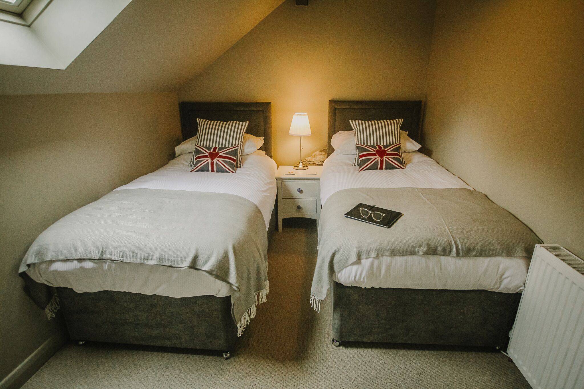 A small, cozy room with two beds, a nightstand with a lamp, and a radiator.