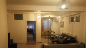 Una sala de estar con sofá, una mesa de centro y un comedor con mesa y sillas. Hay una ventana con cortinas y un equipo de aire acondicionado en la pared.