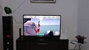 Un televisor de pantalla plana sobre un mueble negro, un cuadro enmarcado encima y una pequeña mesa con una planta en maceta al lado del mueble.