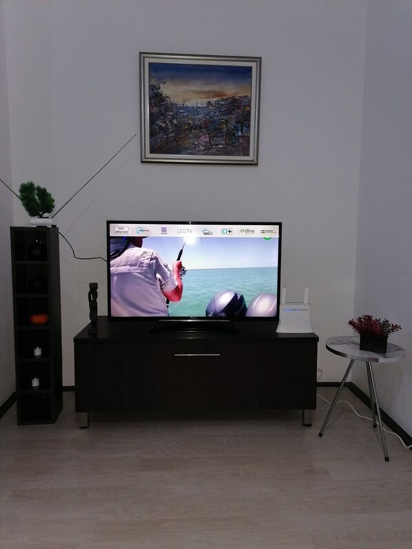 A flat-screen TV on a black entertainment unit, a framed painting above it, and a small table with a potted plant beside the unit.