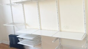 A room with a white metal shelving unit against a plain wall, wooden flooring, and a black radiator. - GREAT UPPER MANHATTAN APARTMENTS FOR GROUP AND FAMILIES (new york)