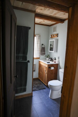 A bathroom with a wooden vanity, a toilet, a towel rack, and a small window with blinds. - Meadow View Cottage (Manheim)