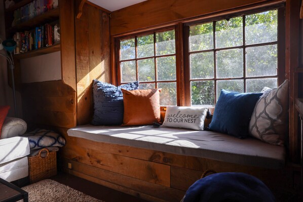 A cozy cabin interior with a built-in bench by a large window, a bookshelf, and a welcome sign. - Meadow View Cottage (Manheim)