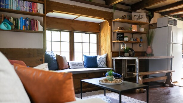A cozy living room with a sofa, a coffee table, and a window seat with cushions. There are bookshelves and a kitchen area visible in the background. - Meadow View Cottage (Manheim)