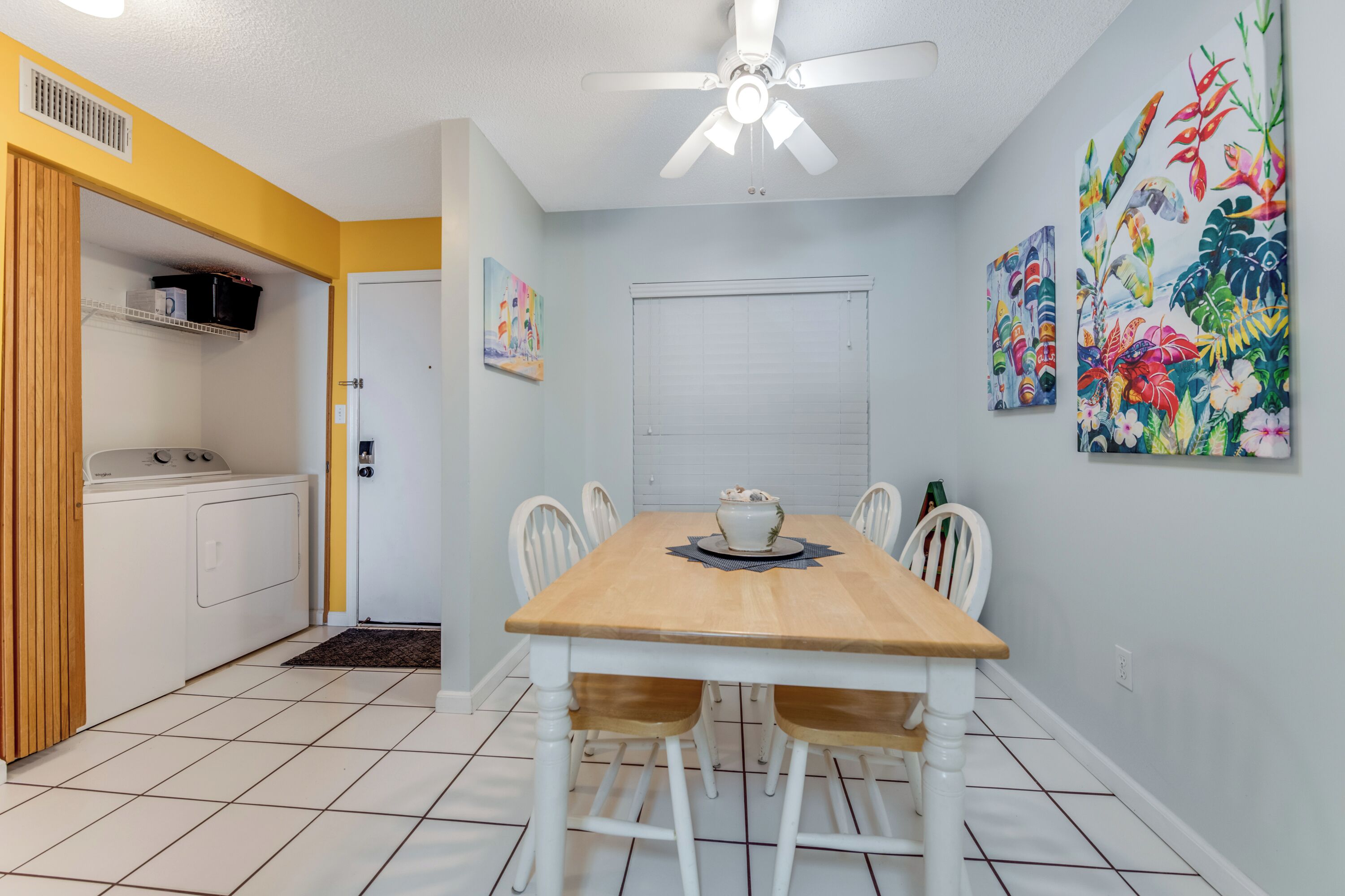 A dining area with a wooden table and chairs, a ceiling fan, and two wall-mounted paintings.