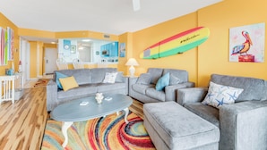 Una sala de estar con ventilador de techo, un tapete colorido y una decoración de tabla de surf en la pared.