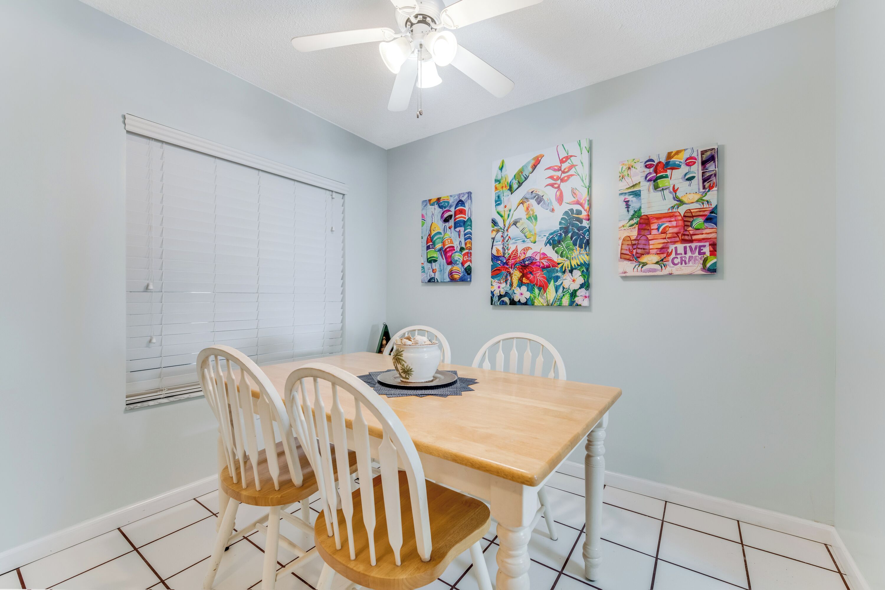 A dining area with a wooden table and white chairs, a ceiling fan, and three colorful wall art pieces.