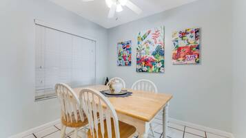 A dining area with a wooden table and white chairs, a ceiling fan, and three colorful wall art pieces.